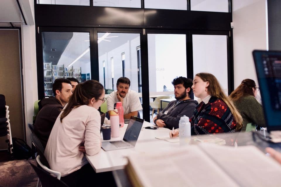 Students working around a table with laptops in a light-filled room.